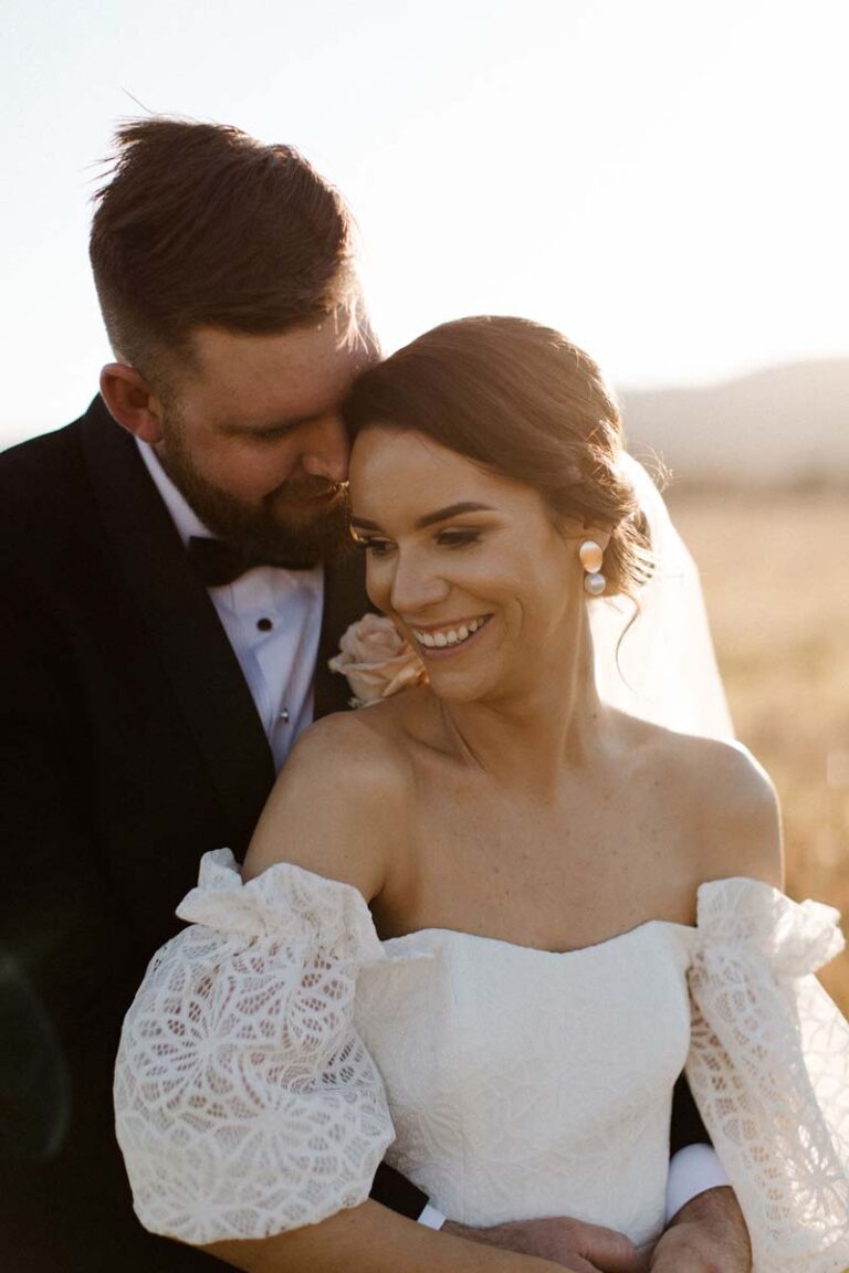 the groom embracing his wife on a field while the sunsets behind them