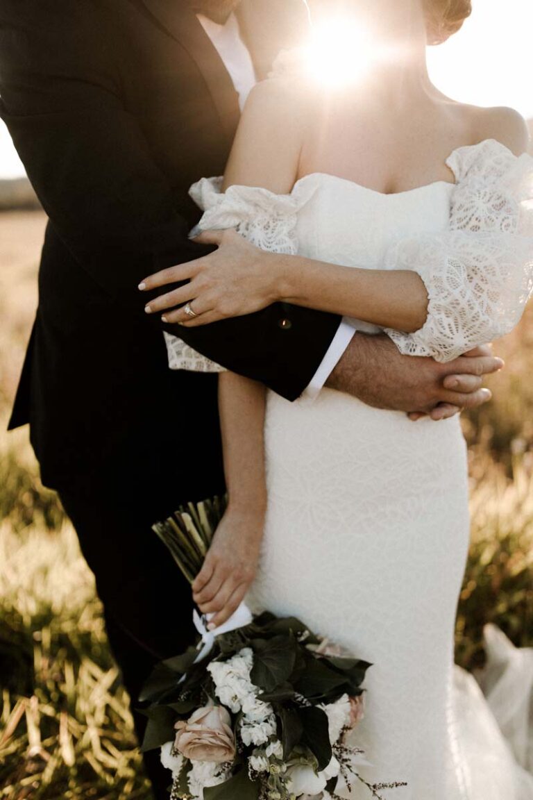 the bride and groom embracing in a field while the sun sets behind them, focusing on the hands and bouquet
