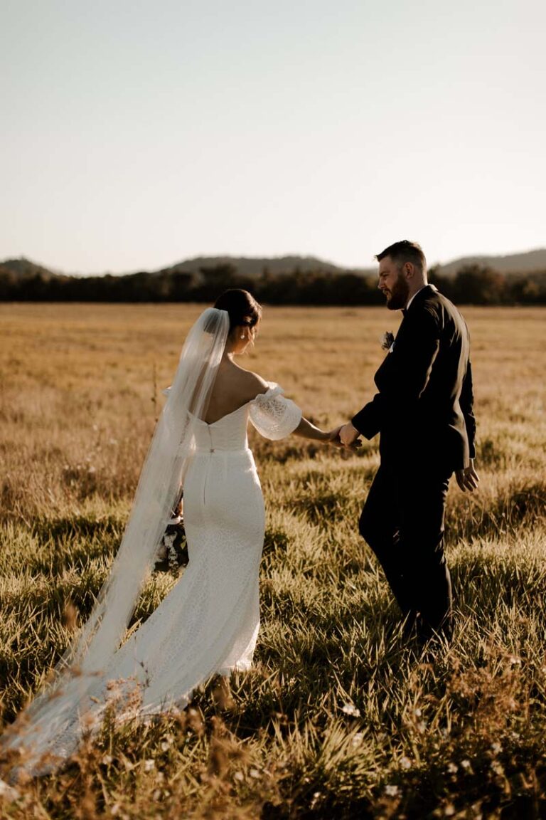 the bride and groom stroll hand in hand through a sunny field, surrounded by tall grass and wildflowers