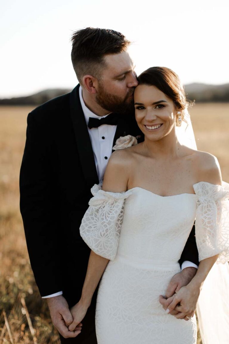 the bride and groom standing together in a sunny field, surrounded by green grass and wildflowers, smiling happily