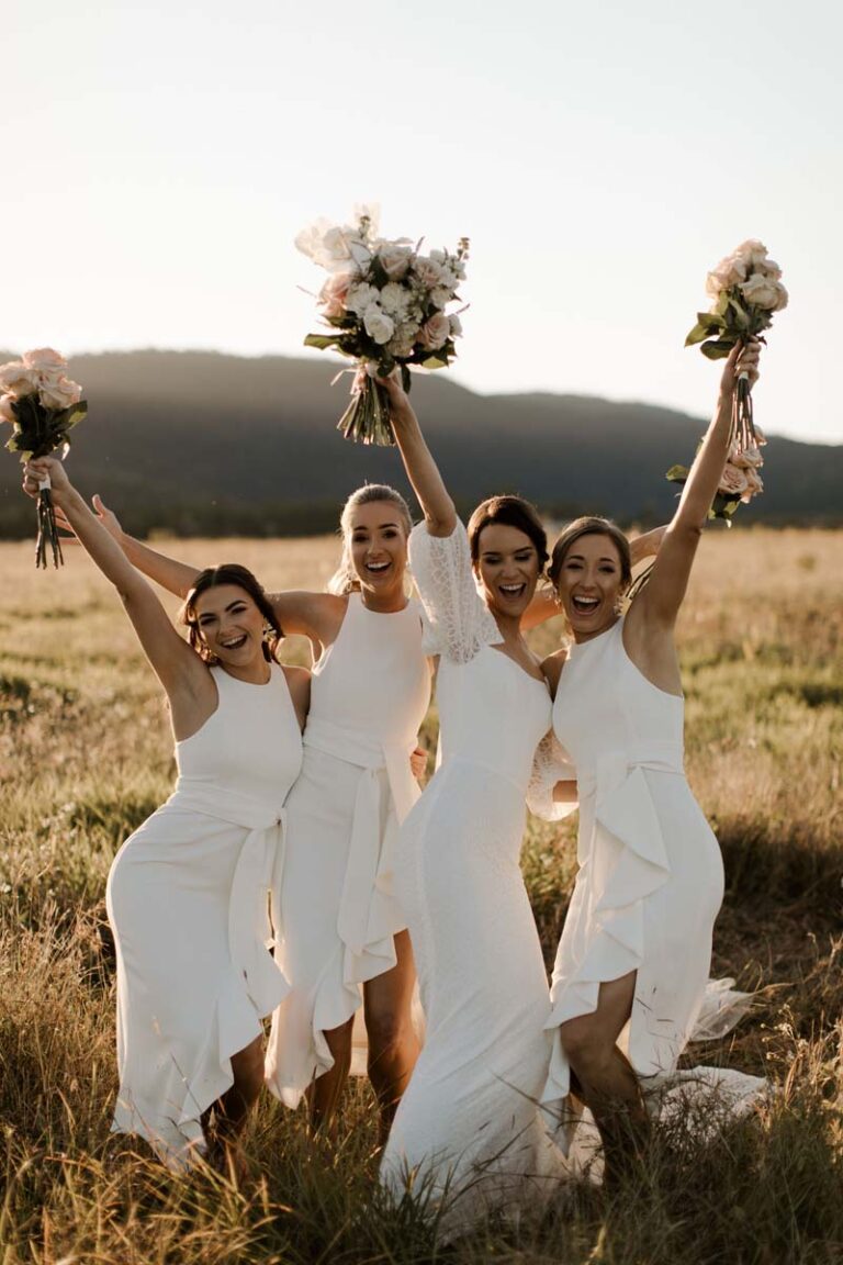 four joyful bridesmaids in white wedding dresses holding bouquets in a sunny field.