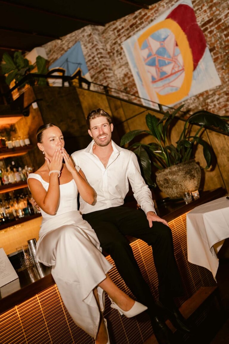 a bride and groom seated on stools, the bride is blowing a kiss