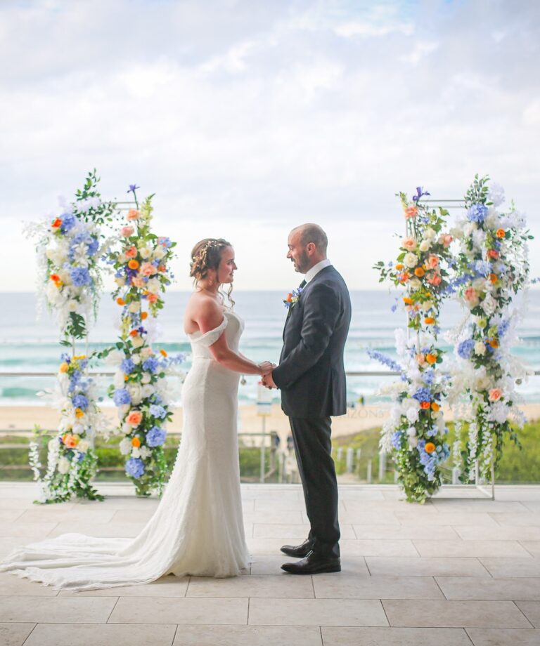 A couple standing between two floral decorations, holding hands