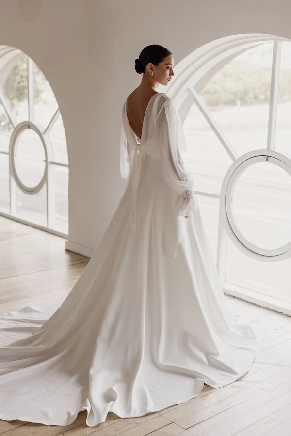 A bride looking out a window in a long sleeve wedding gown with sheer sleeves