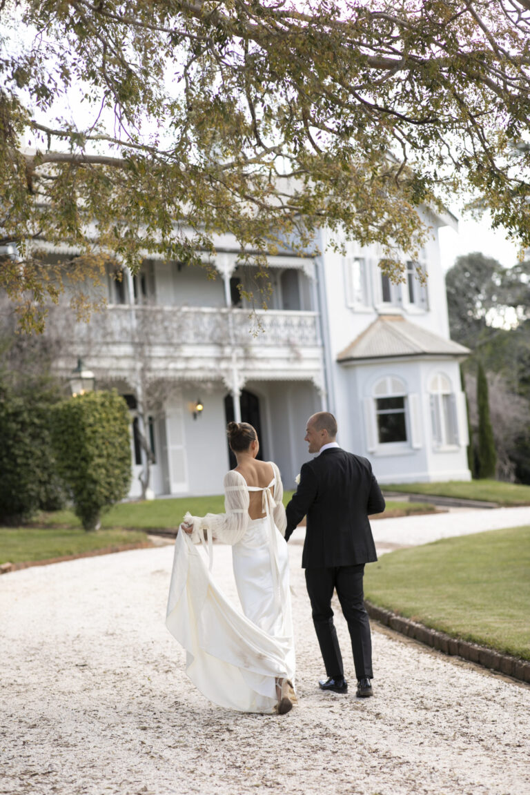 A bride and groom walking down the driveway in front of a large house.