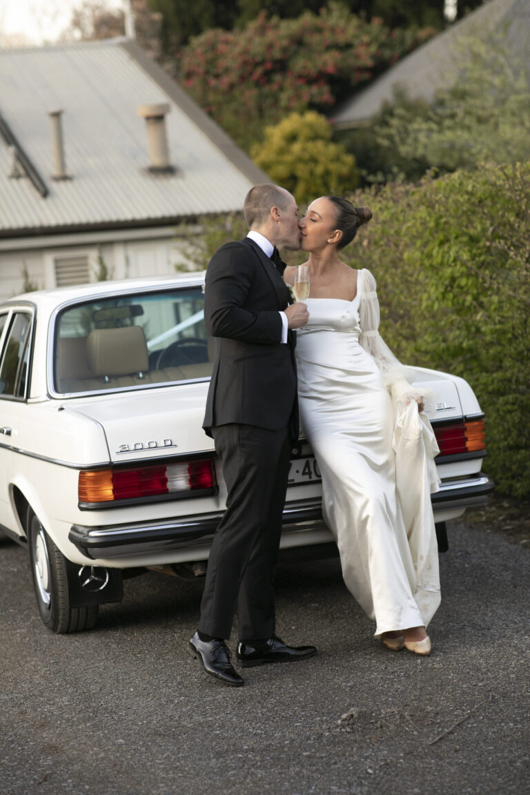 The bride and groom kissing in front of a white car.