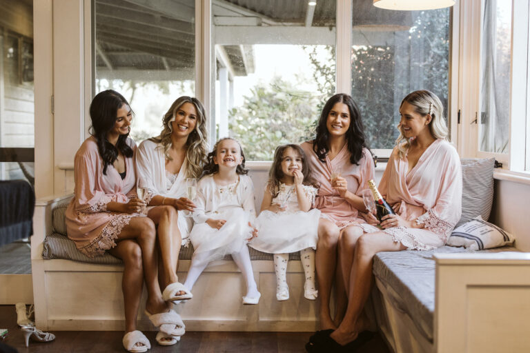 Bridesmaids and flower girl in pink robes sitting on a couch at a wedding venue.