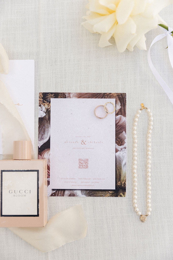 A flower, perfume, a necklace, rings on top of a wedding invitation.