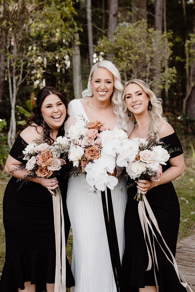 A bride an two bridesmaids in black dresses holding bouquets, standing together at a wedding.