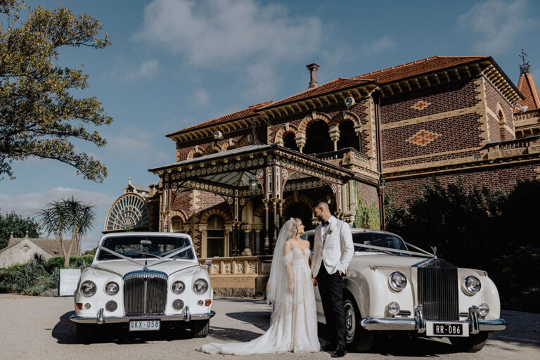 Couple posing in front of two luxury car and a mansion.
