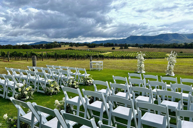 A beautiful outdoor wedding ceremony in a vineyard with rows of grapevines, white chairs, and a floral arch.