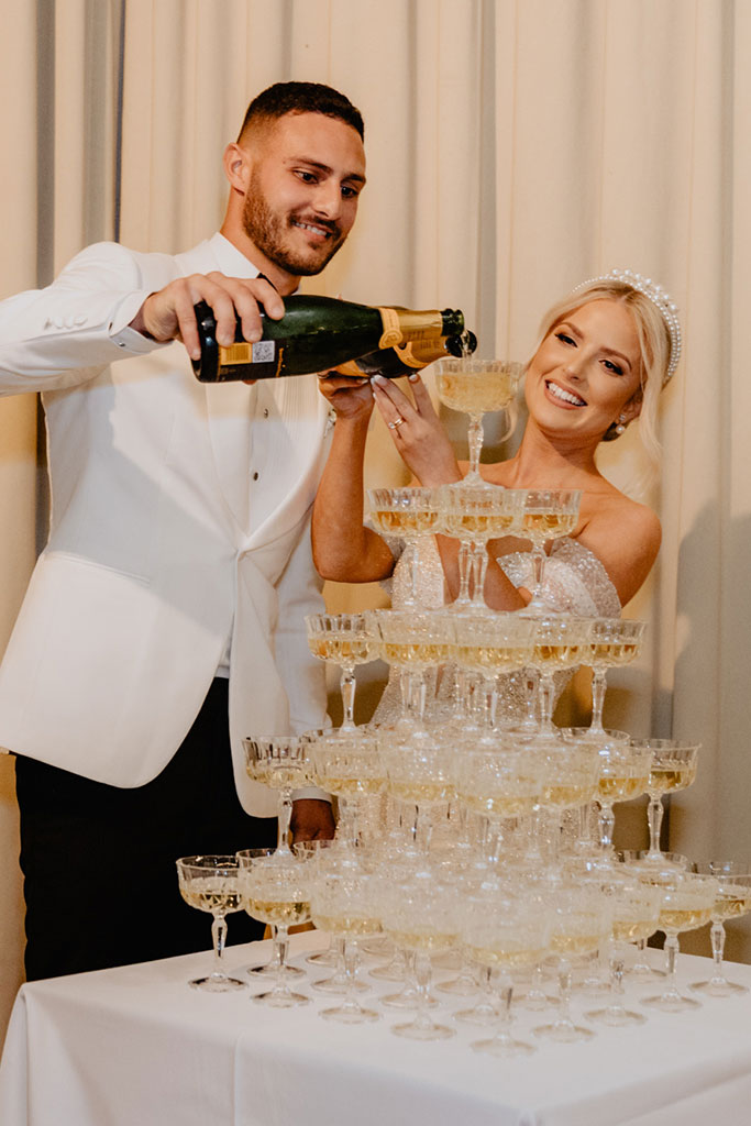 A couple pouring champagne on a tower of champagne glasses.