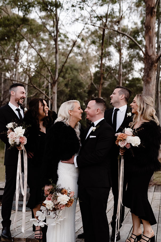 A couple smiling at each other with people celebrating behind them.
