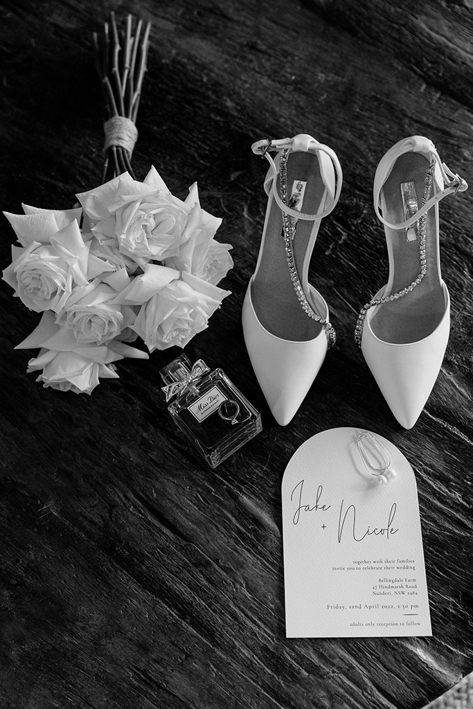 A black and white photo of a wedding shoes, flowers, perfume and a wedding invitation.