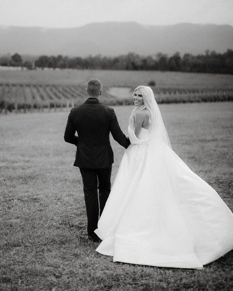 A bride and groom strolling through a vineyard, hand in hand, surrounded by lush green vines.