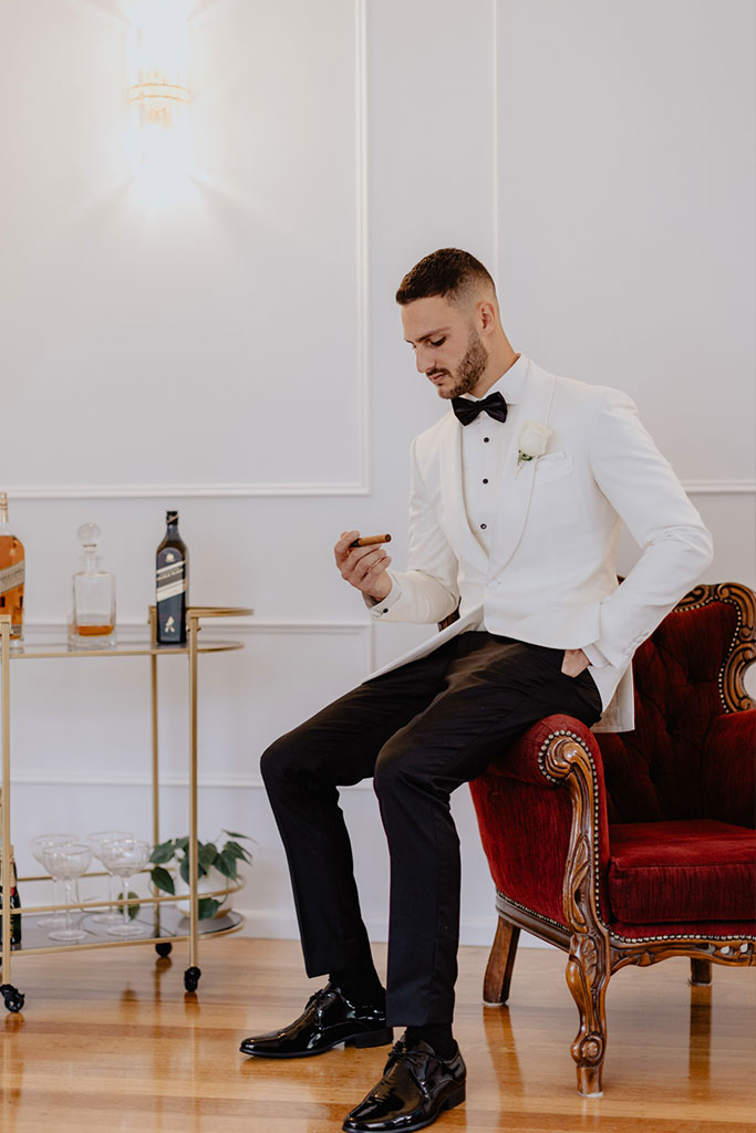 A man in a tuxedo sitting on a chair holding a cigarette.