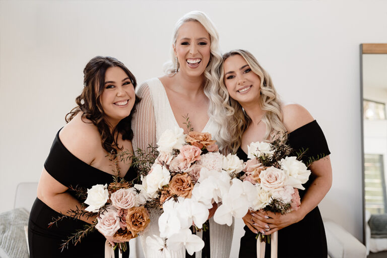 A bride and two women beside her wearing black dresses, each holding a bouquet of flowers..