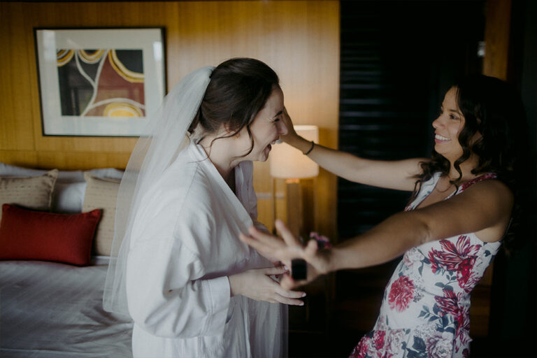 A bride in a white gown, smiling as a woman hugs her.