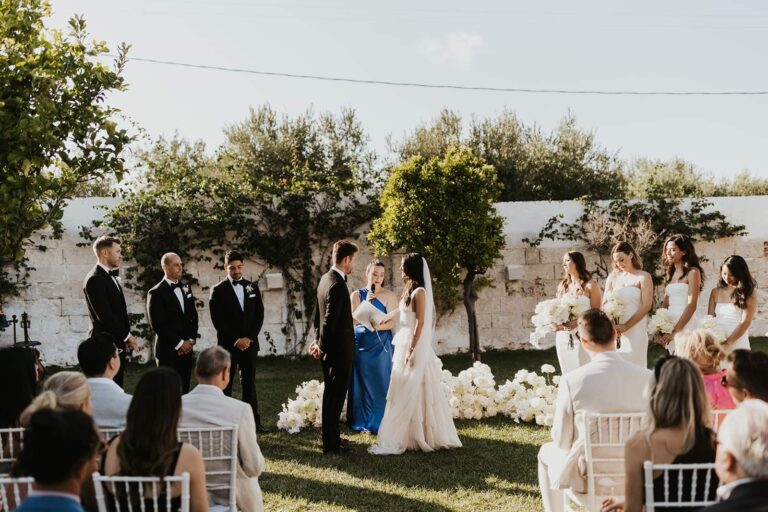 A wedding ceremony in a garden with guests seated on white chairs, surrounded by blooming flowers and greenery.