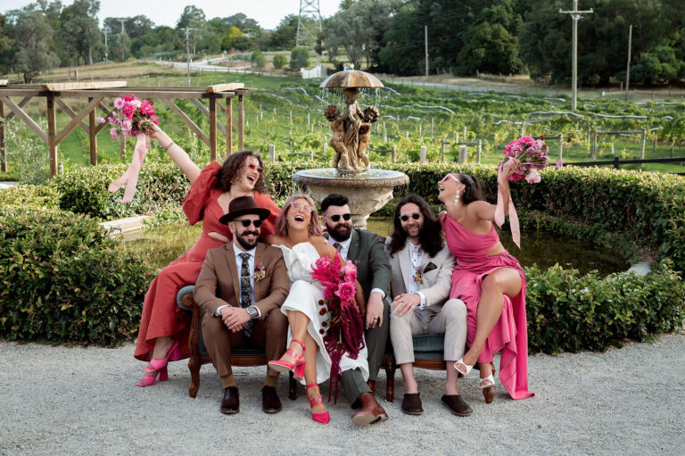 Bride, groom, bridesmaids and groomsmen laughing together on a wooden sofa by the fountain at the best wedding venue in Yarra Valley.