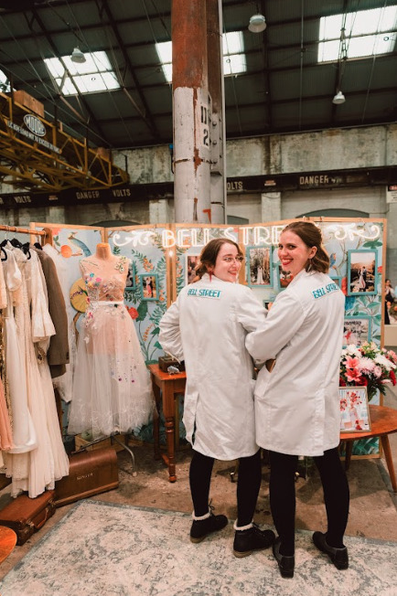 Two women in lab coats standing in front of a display of dresses.
