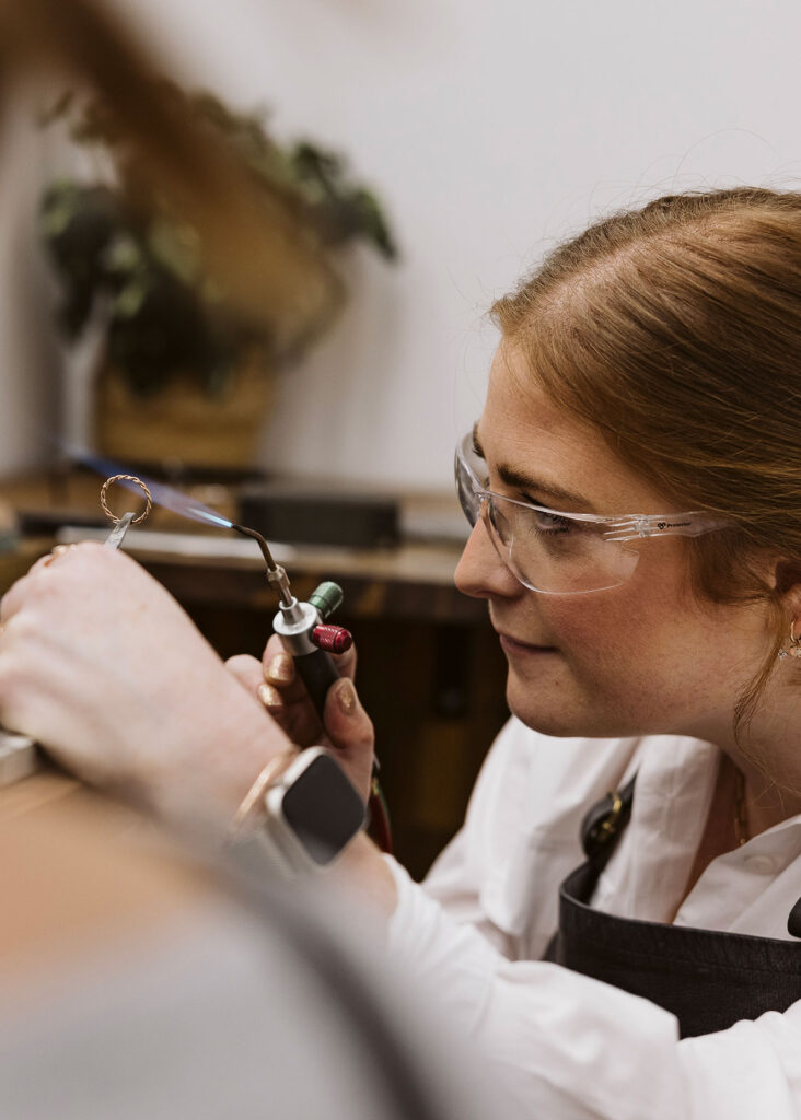 Meg Maskell crafting custom engagement rings in her workshop, torching a gold ring to smooth its surface.