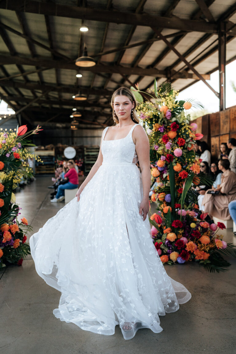 A bride in a white wedding gown posing in front of a colorful array of flowers.