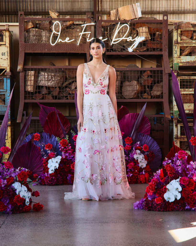Woman posing in a deep V-neck sleeveless wedding dress with floral detail pattern at a bridal runway show in Melbourne.