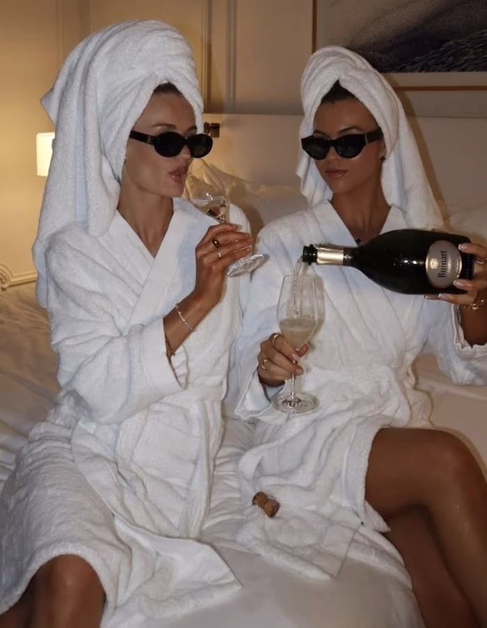 Two women in robes and white towels wrapped around their hair wearing sunglasses and drinking champagne at a hens party