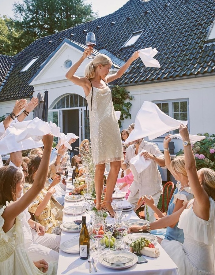 A woman in a sequinned dress dancing on a lunch table as her friends cheer her on