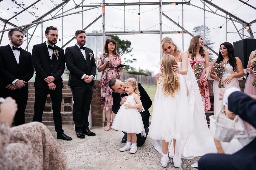 A bride, groom, and a little girl share a kiss in front of a crowd of people at their wedding.