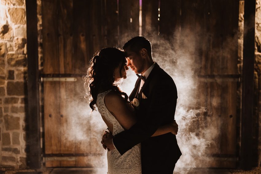 Wedding photo at rustic barn on ranch with beautiful scenery, bride and groom embracing.