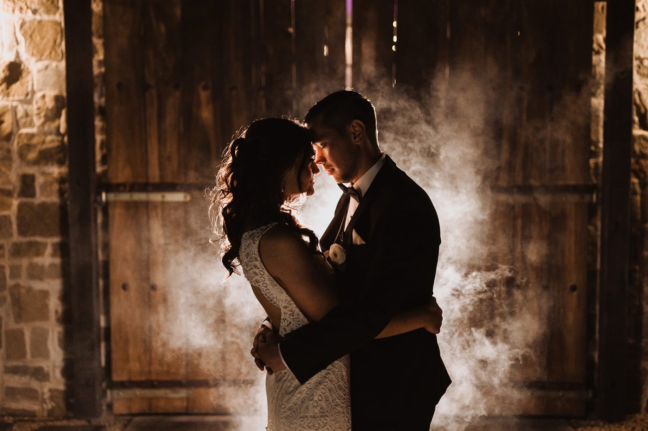 Wedding photo at rustic barn on ranch with beautiful scenery, bride and groom embracing.