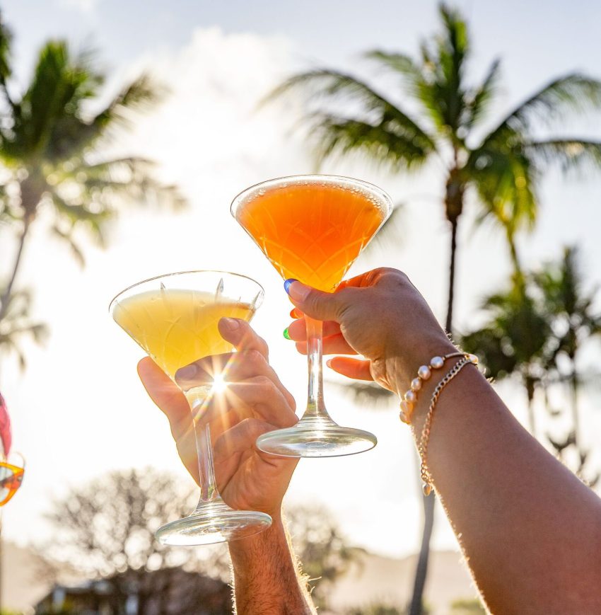 Two people raising glasses in front of palm trees, celebrating with drinks.